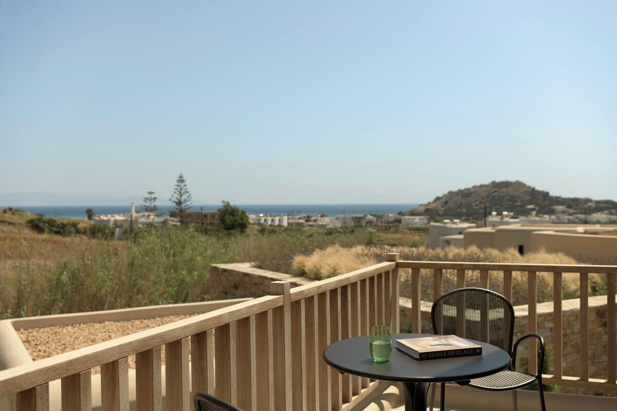 A balcony with a table and chair, overlooking a coastal landscape with buildings and vegetation.