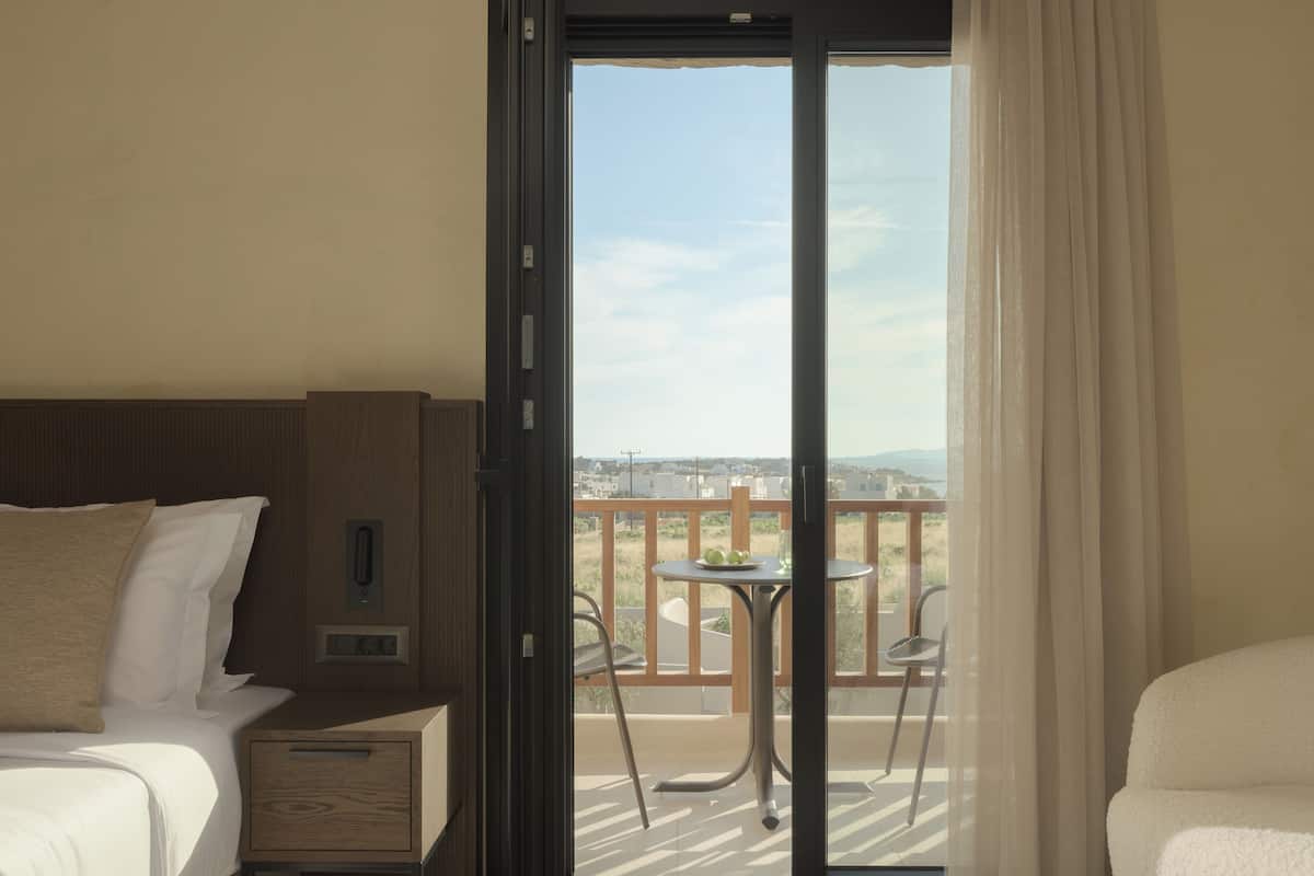A hotel room with a bed, a nightstand, a window with a view of the balcony, and a sliding door.