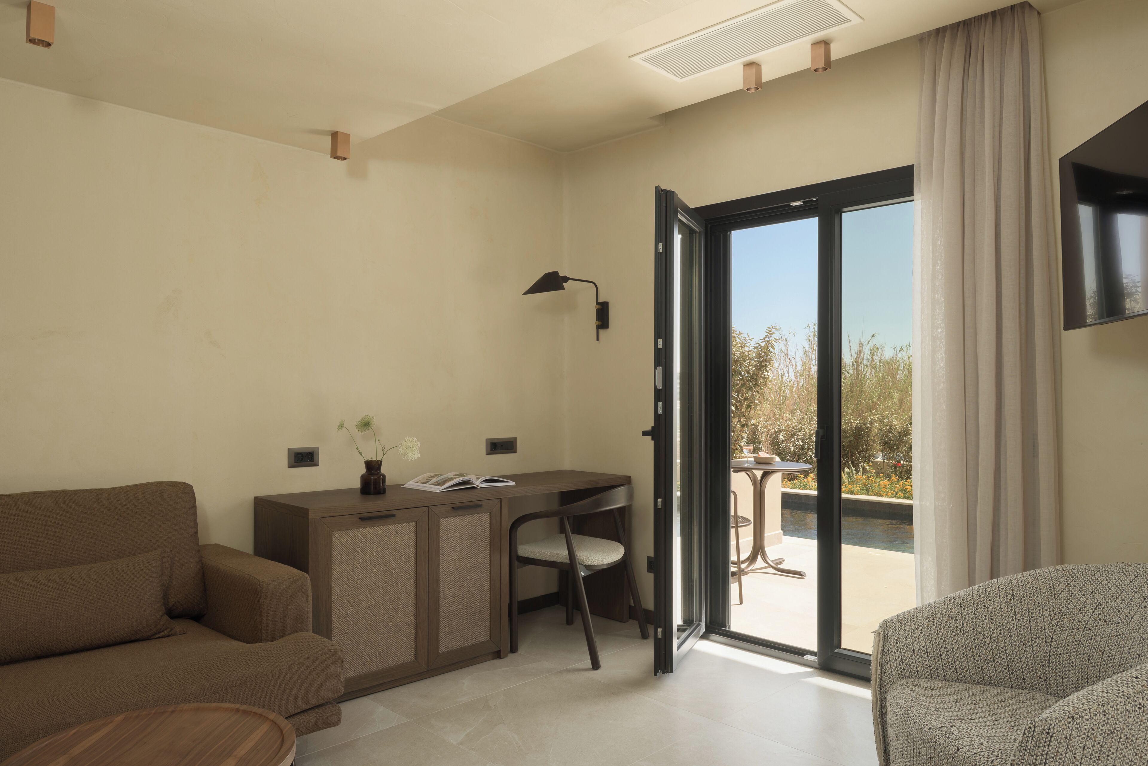 A modern hotel room with a sofa, a desk with a lamp, and a view of the outdoors through a sliding door.