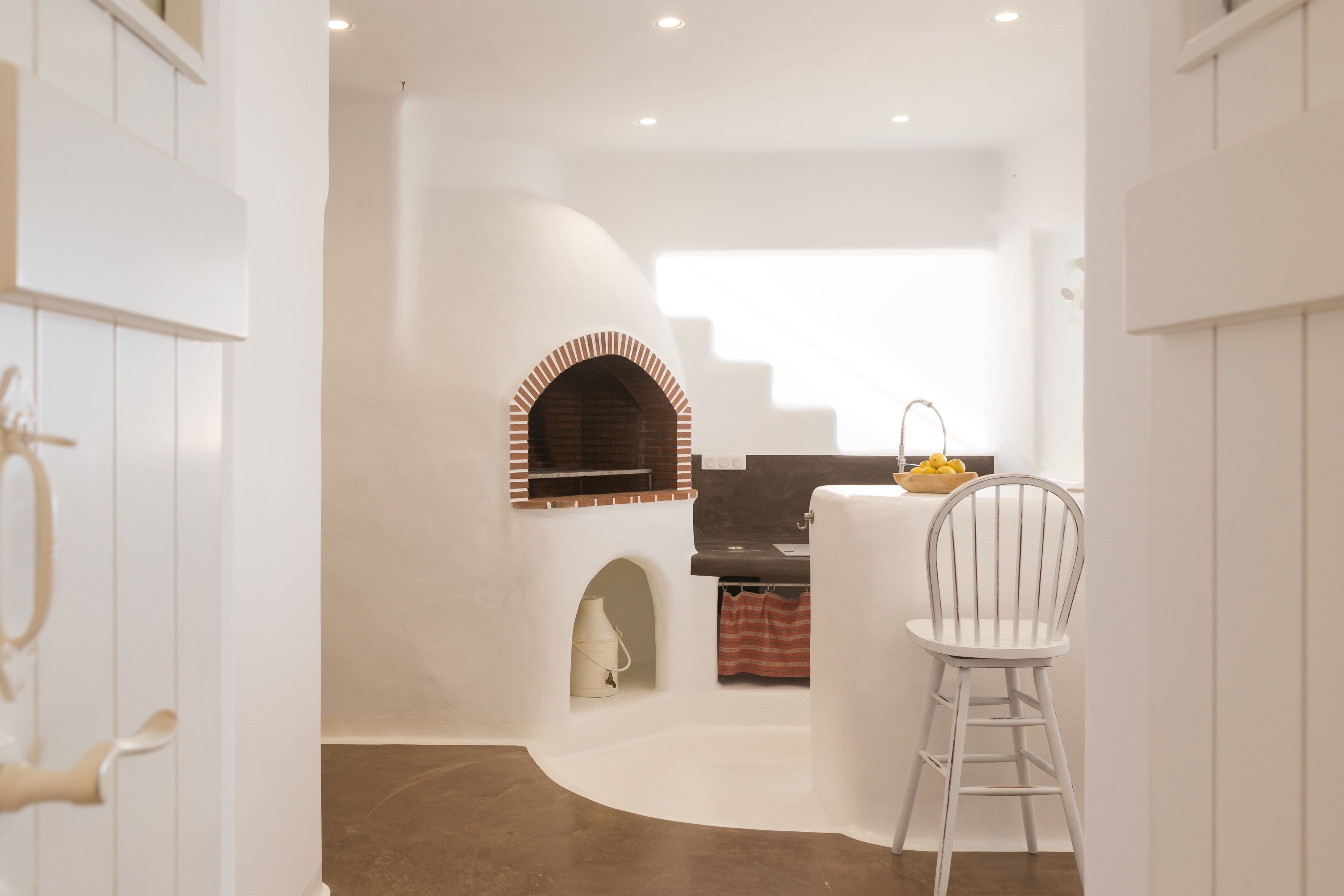 A modern kitchen with a brick oven, white cabinetry, and a dining area with a white chair.