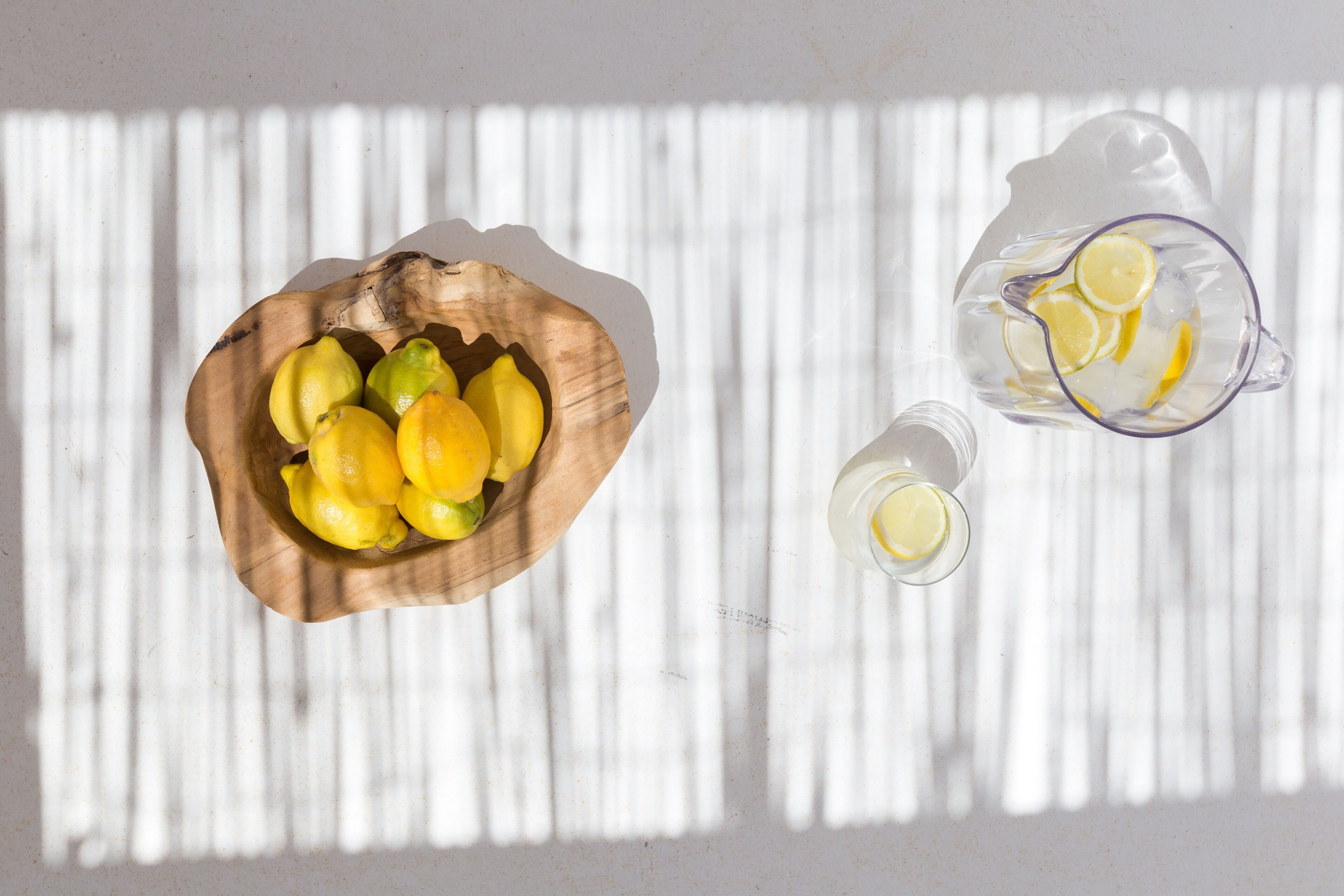 A wooden bowl filled with lemons and a pitcher with lemon slices on a white surface with vertical shadows.