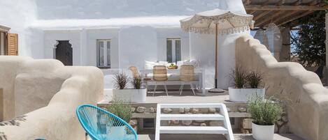 A white-walled building with a patio area featuring wicker chairs, a bench, and potted plants.