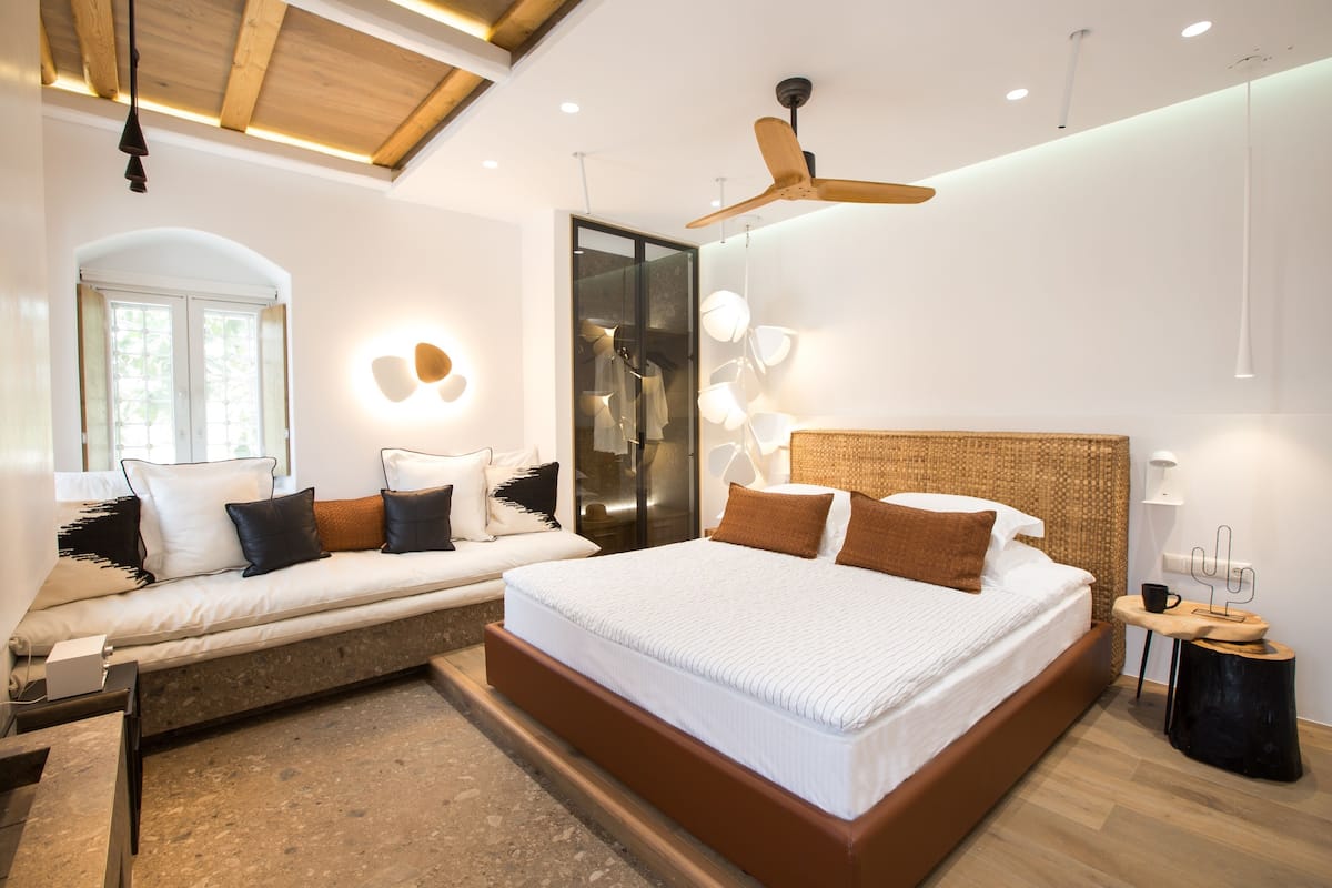 A modern bedroom with a large bed, a seating area, a ceiling fan, and a large window.