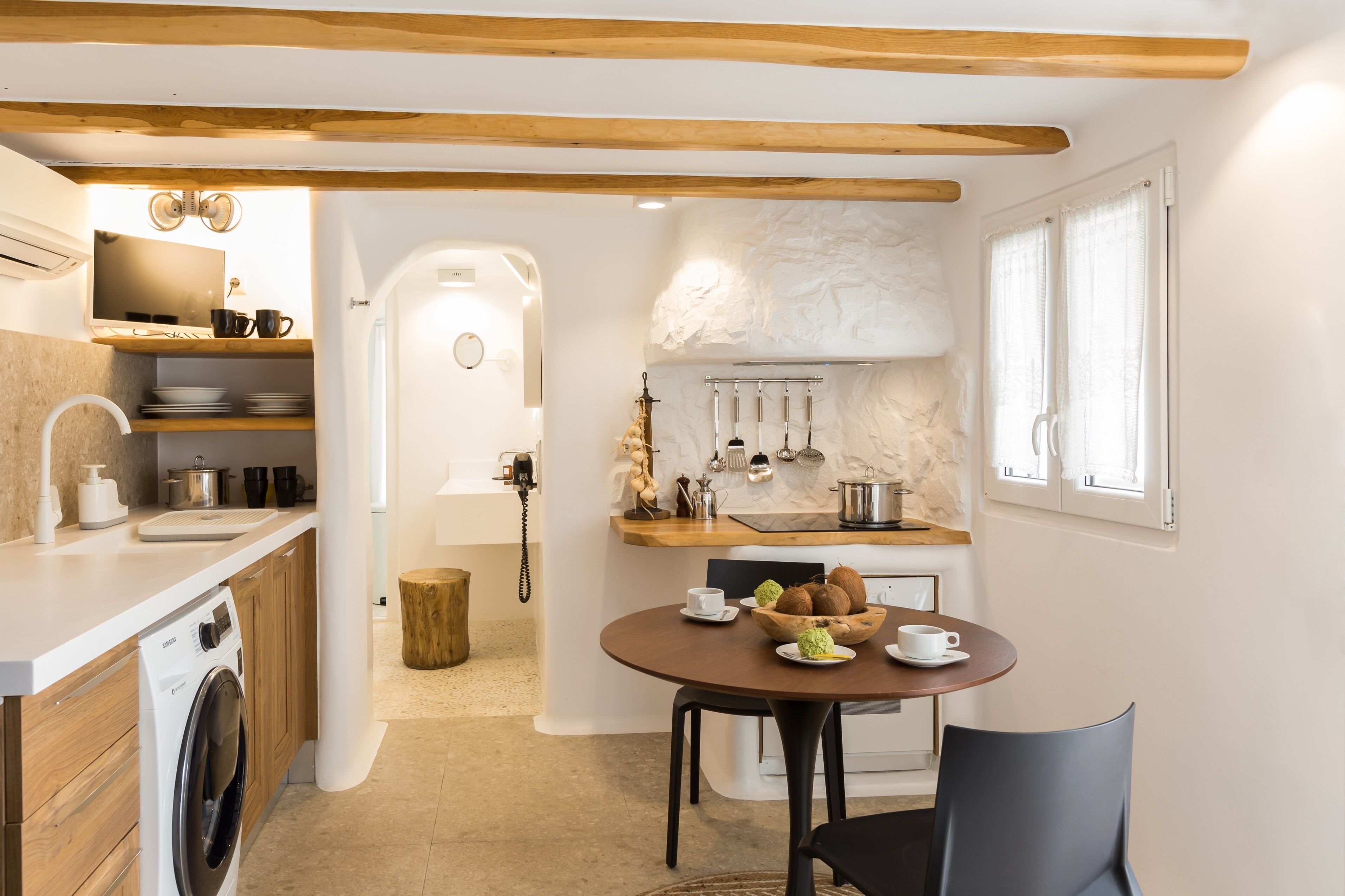 A compact kitchen with wooden beams, a washing machine, a dining table with a bowl of fruit, and a coffee cup.