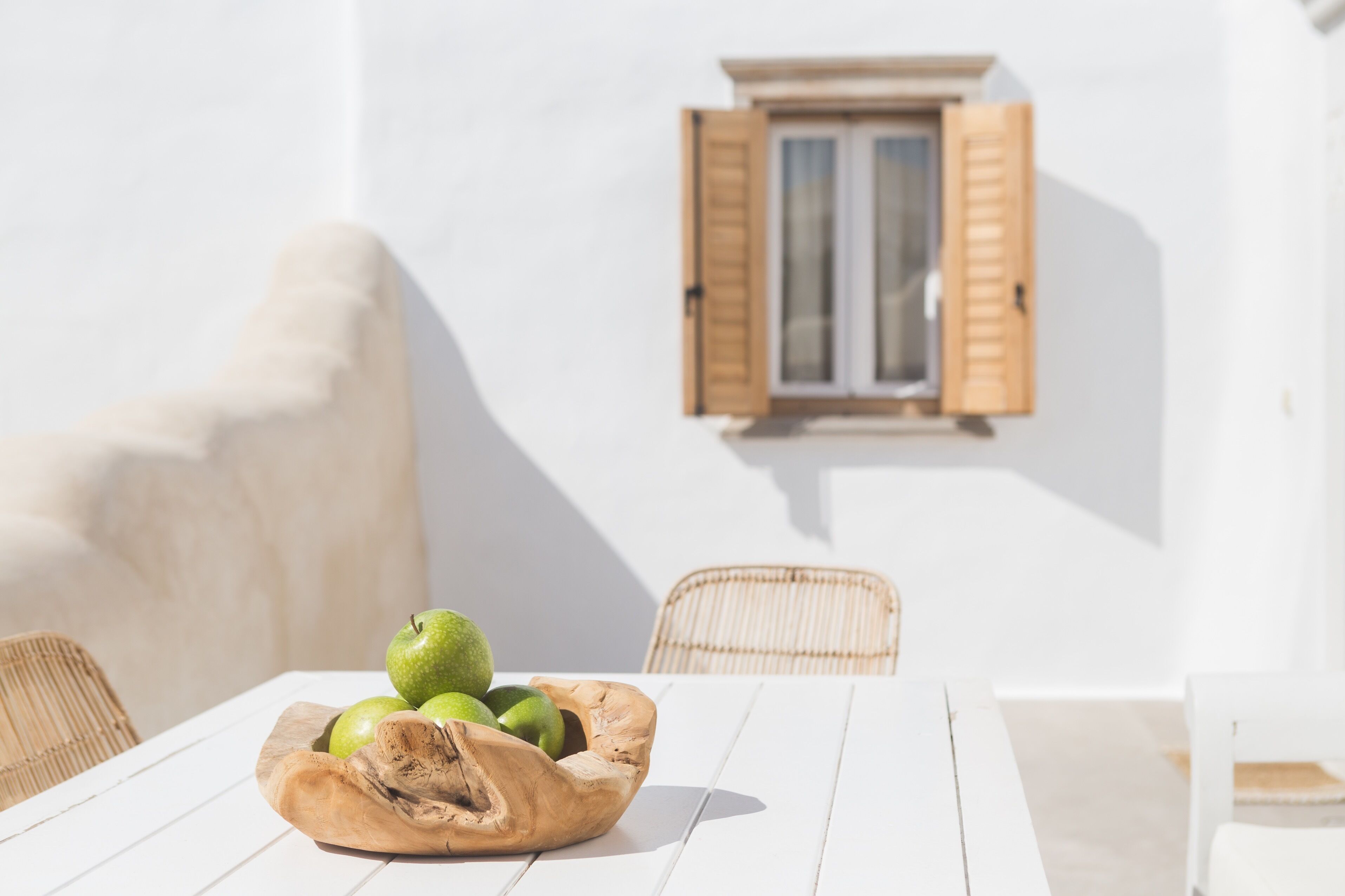 A white table with a wooden bowl containing green apples and a pear, set against a white wall with a window and wooden shutters.