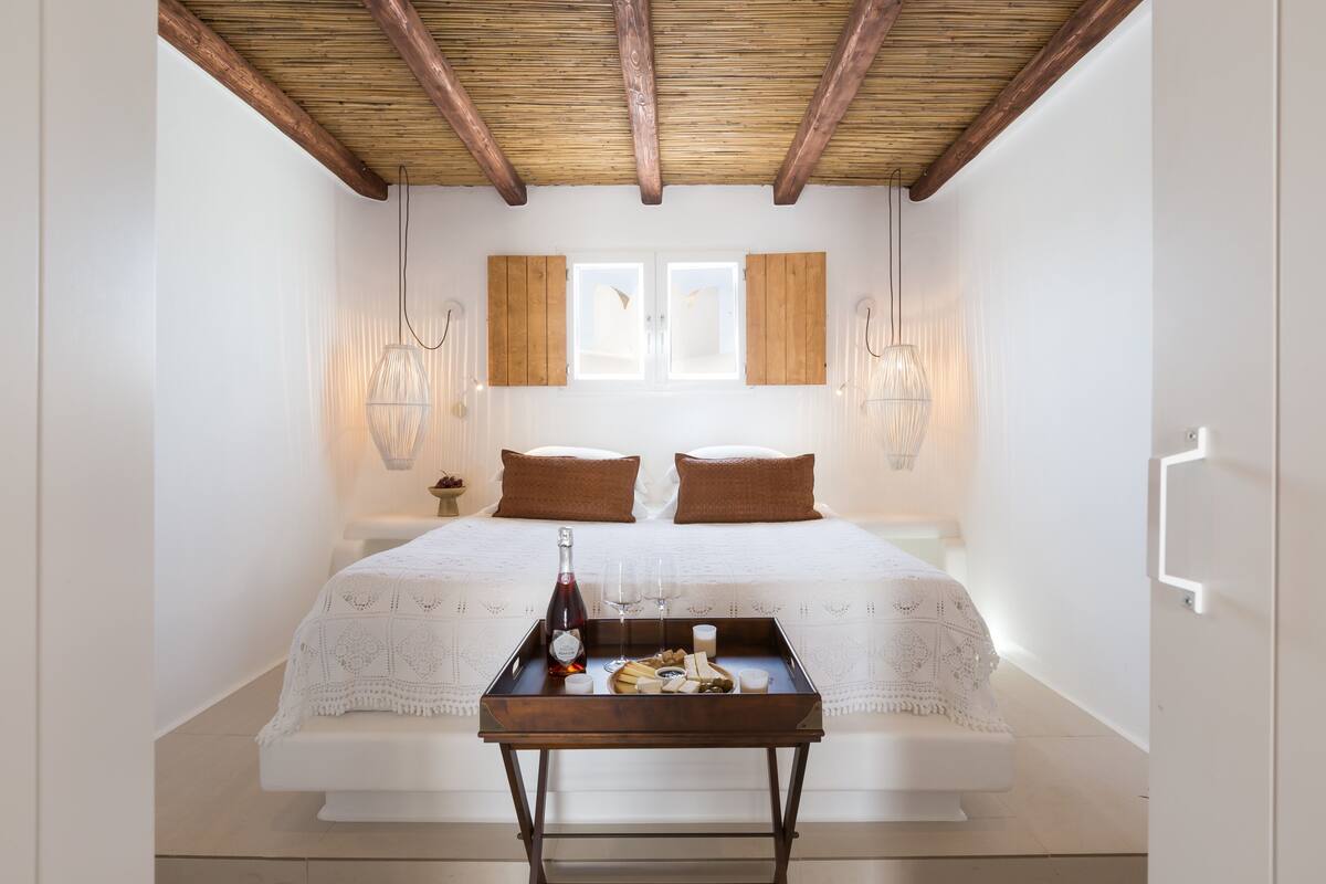 A bedroom with a wooden ceiling, a bed with a white bedspread, two brown pillows, a bedside table with a tray of food and drinks, and two wall-mounted lamps.
