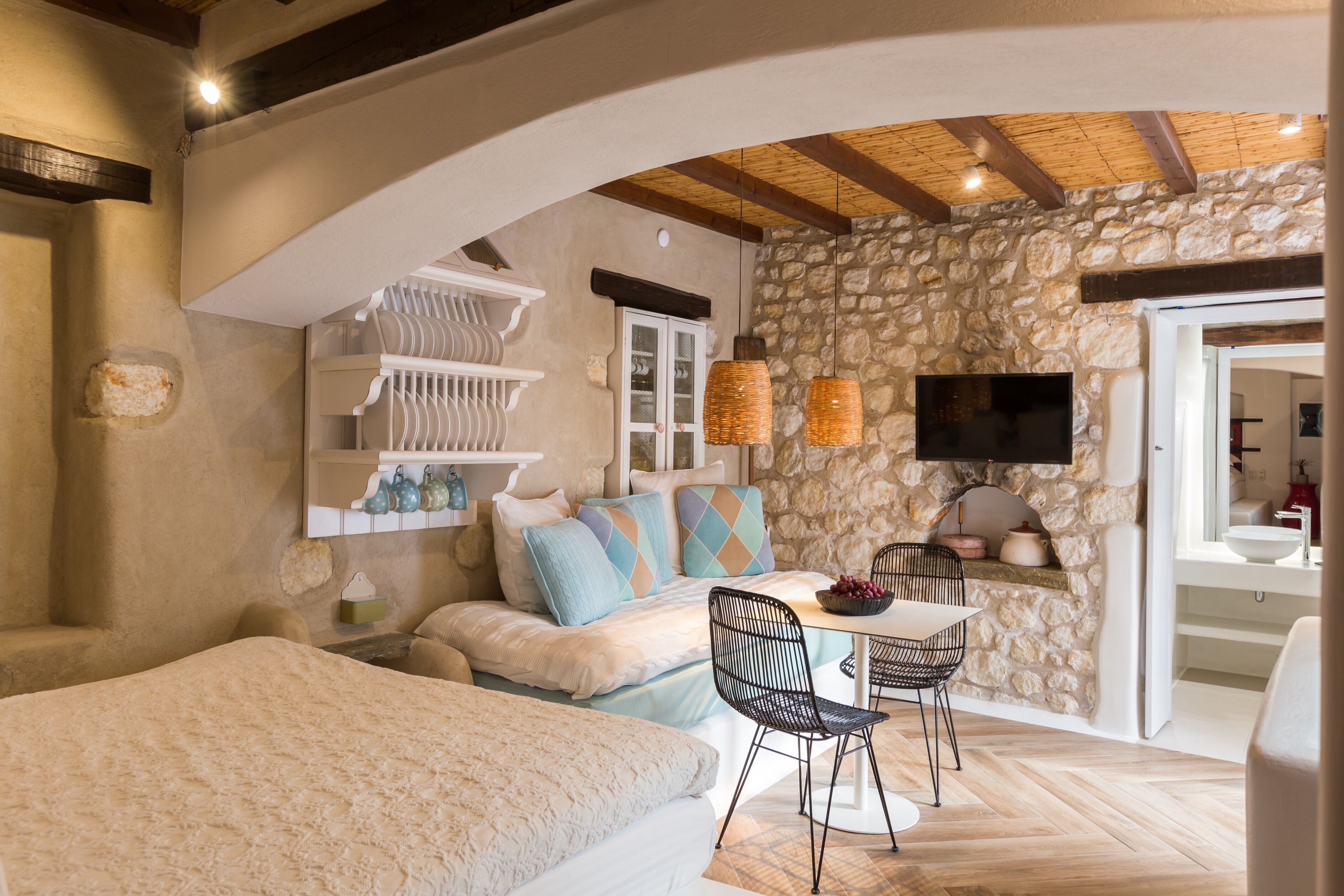 A cozy bedroom with a large bed, a dining area with a table and chairs, a TV mounted on a stone wall, and a staircase leading to an upper level.