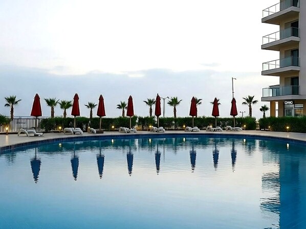 A swimming pool with lounge chairs and umbrellas, surrounded by palm trees and a building with balconies. - Luxurious Chalet in El Manasra, Egypt - Your Home Away From Home (Al-Manasrah)