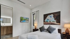 A hotel room with a bed, a nightstand with a lamp, a painting of a beach scene, a bathroom with a mirror, and a view of the outdoors. - Miami Bali Villa Near Design District with Pool and Spa (Miami)