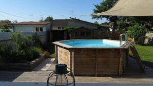 Ein hölzernes Whirlpool mit einer Leiter, umgeben von einem Garten mit einer Holzterrasse, einem kleinen Hocker und einer großen Markise.