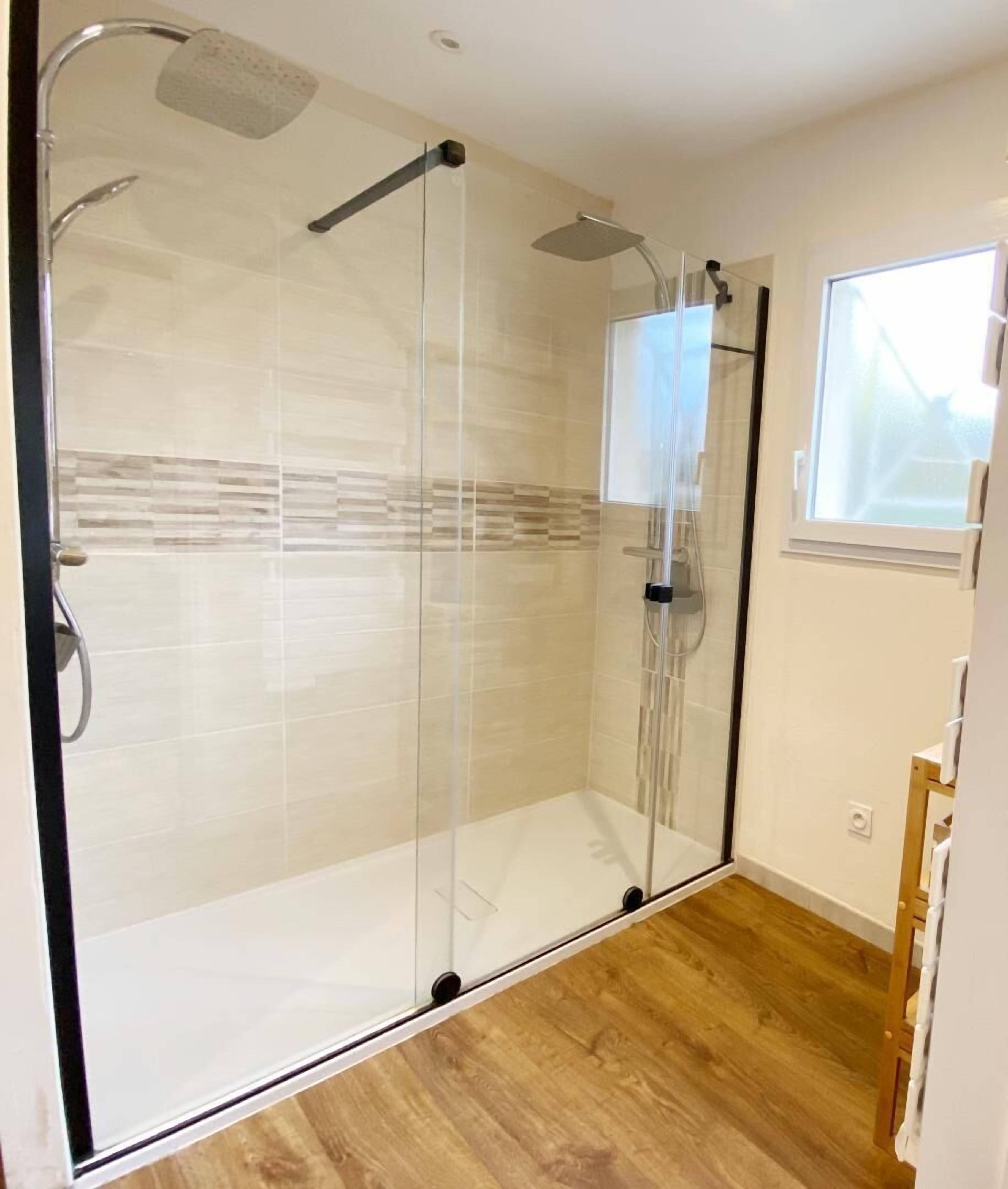 A glass-enclosed shower with a handheld showerhead and a fixed showerhead, tiled walls, and a wooden floor.