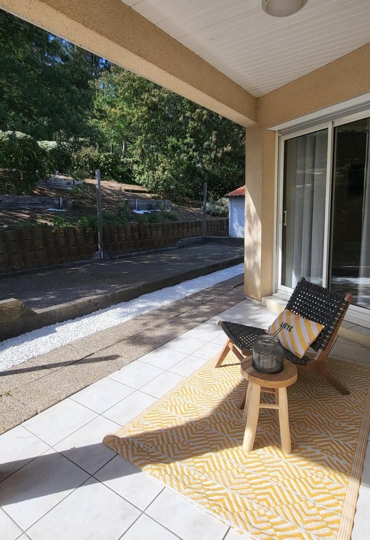 A covered patio with a chair and a small stool, a patterned rug, and a view of a landscaped area with trees and a building in the background.