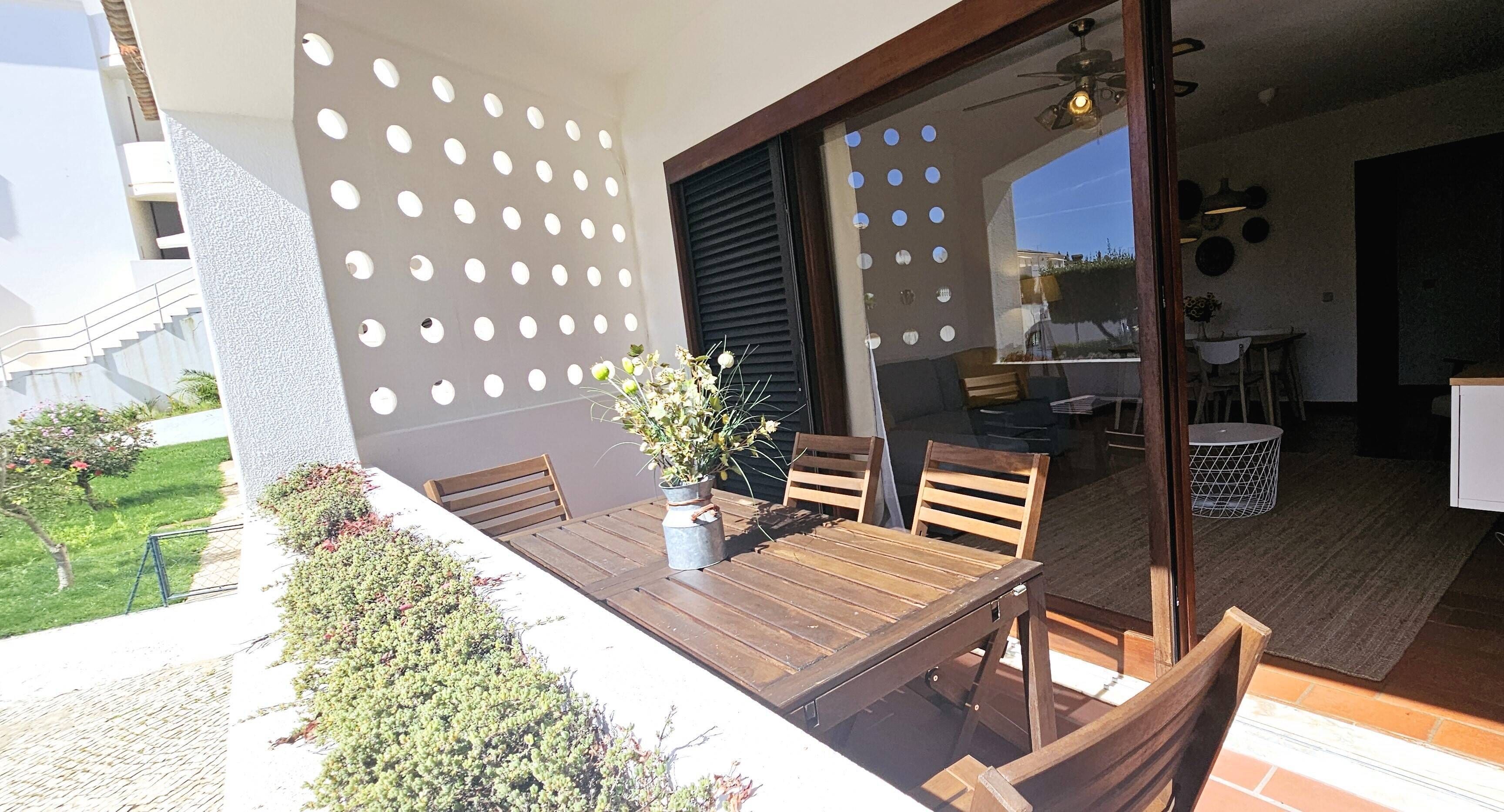 An outdoor patio with a wooden table and chairs, a potted plant, and a wall with circular cutouts.