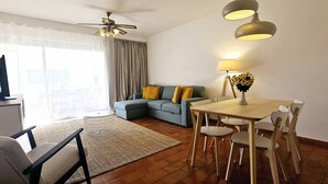 A modern living room with a sofa, a dining table with chairs, a ceiling fan, and a lamp.