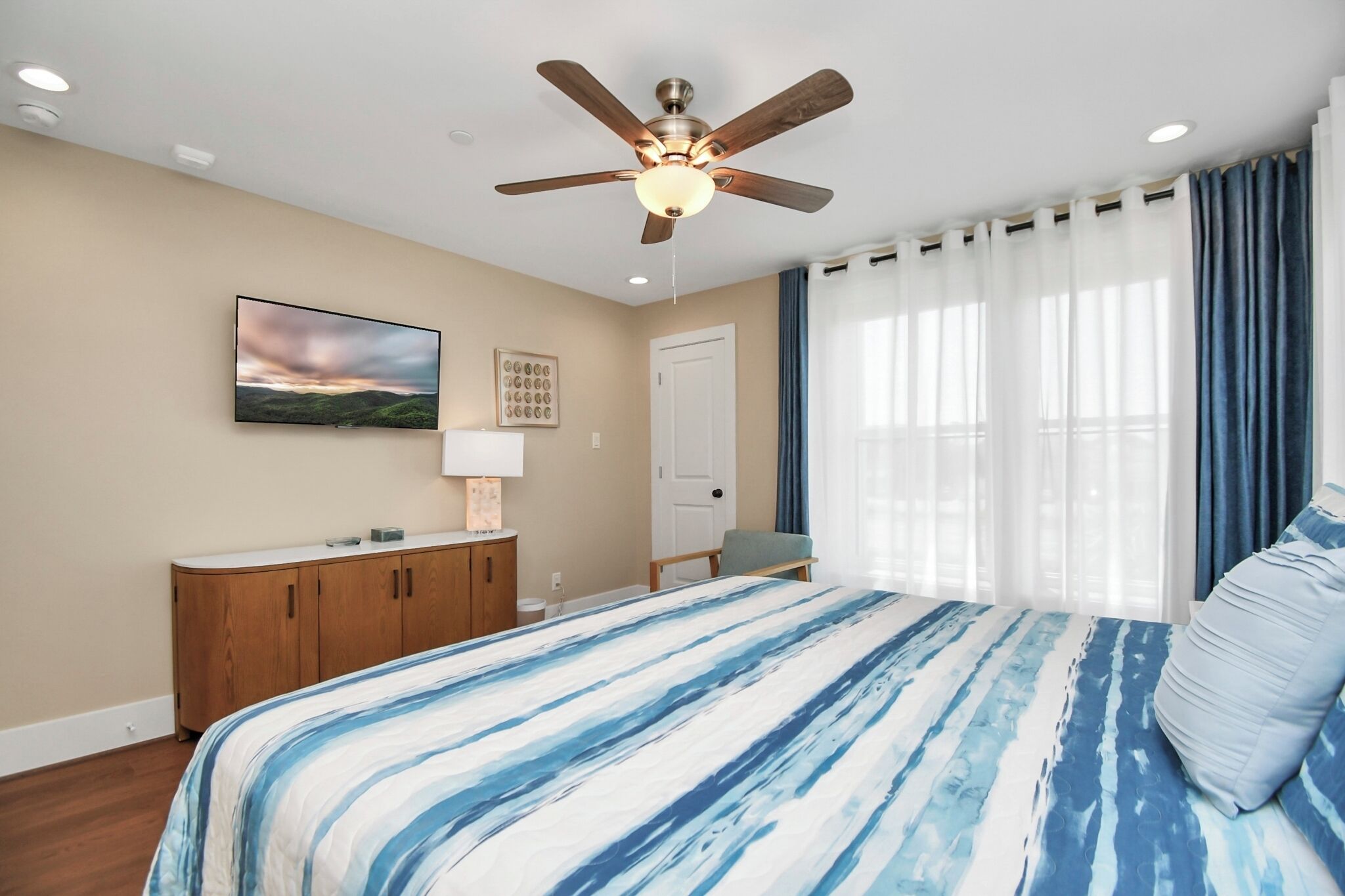 A bedroom with a bed, a ceiling fan, a TV, a dresser, and a window with curtains.