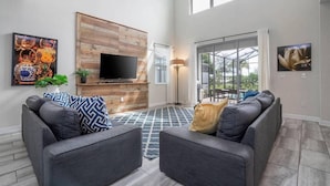 A modern living room with a large TV, grey sofas, patterned cushions, and a wooden accent wall. - Amazing Home at Storey Lake 2 Minutes to Clubhouse (Kissimmee)