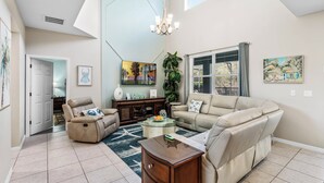A spacious living room with a high ceiling, a chandelier, a beige sofa, a recliner, a coffee table, and a TV on a stand.