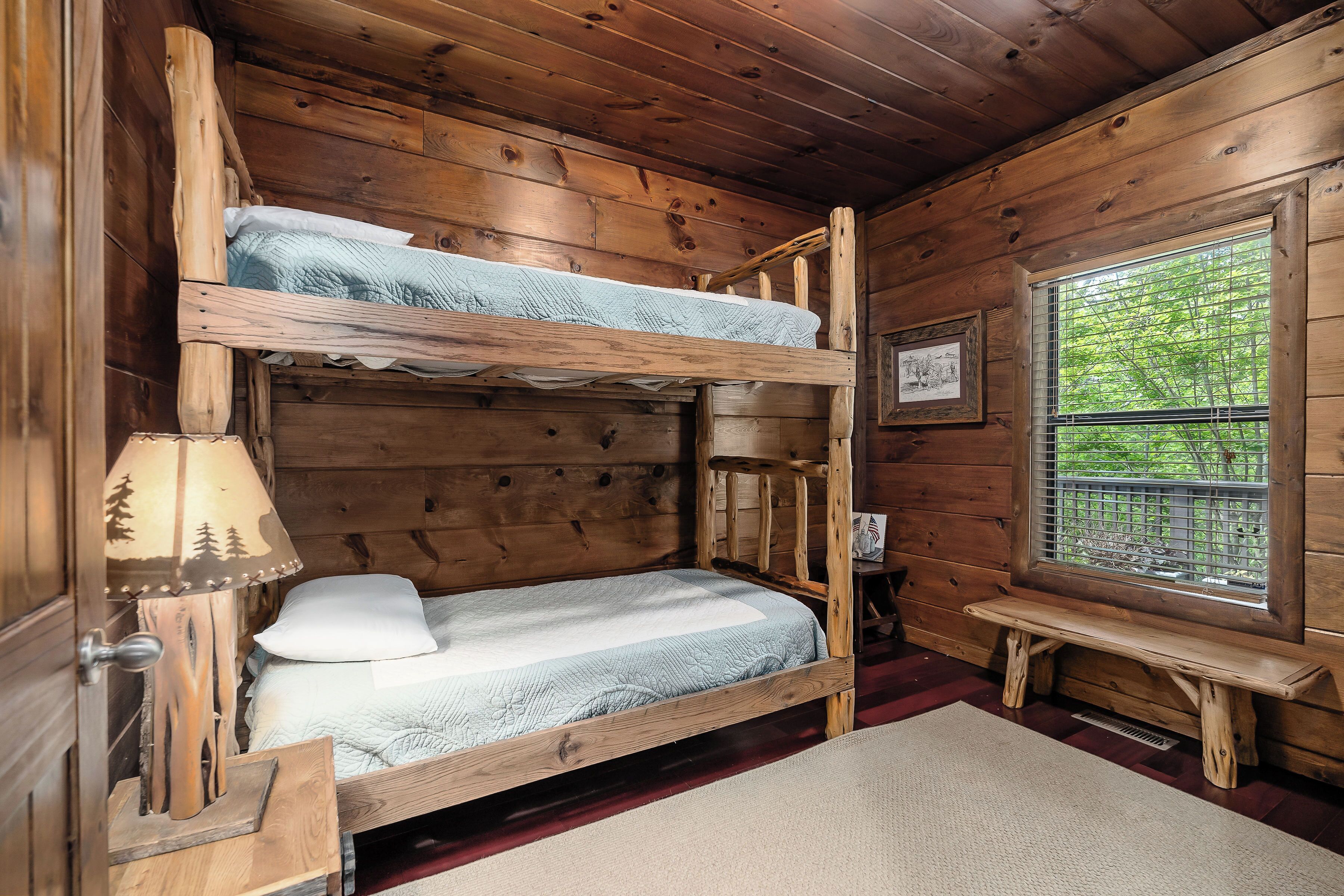 Ein Zimmer einer Holzhütte mit einem Etagenbett, einer Lampe, einem Fenster mit Blick auf Bäume und einem gerahmten Bild an der Wand.