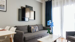 Ein modernes Wohnzimmer mit einem grauen Sofa, einem weißen Couchtisch und einem Esstisch mit Stühlen. An der Wand hängt ein gerahmtes Bild und auf dem Couchtisch steht ein Blumentopf mit blauen Blumen.