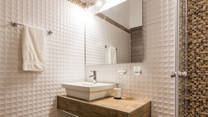 A bathroom with a white tiled wall, a wooden vanity, a white sink, and a mirror.