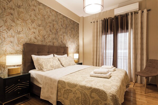 Un lit bien fait, recouvert d’une couvre-lit à motifs, une lampe de chevet et une chaise dans une pièce dont le papier peint est orné de motifs floraux et qui possède une grande fenêtre avec des rideaux.