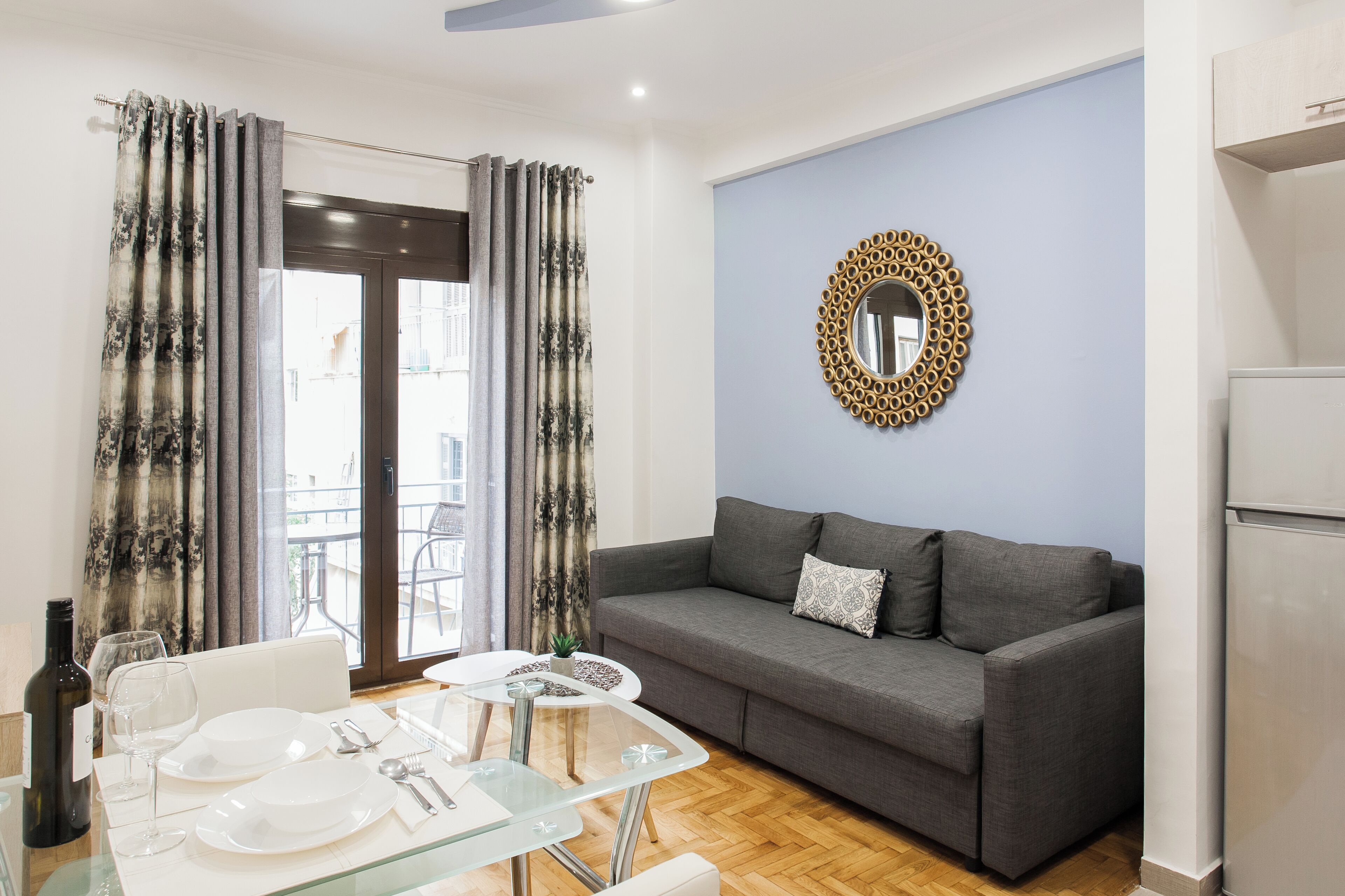 A modern living room with a grey sofa, a glass dining table set for two, and a large window with curtains.