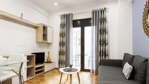 Ett modernt vardagsrum med en grå soffa, ett soffbord i glas och en tv-apparat som står på ett träställ. Det finns en liten växt på bordet och en trähylla ovanför tv:n.