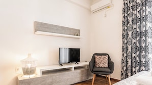 Une chambre d’hôtel moderne avec un téléviseur à écran plat posé sur une console blanche, un fauteuil gris agrémenté d’un coussin à motifs et un lit avec des serviettes blanches soigneusement pliées.