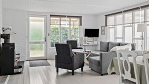 A modern living room with a grey sofa, a dark armchair, a white dining table, and a TV on the wall.