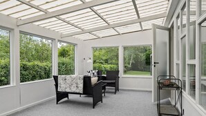 A conservatory with a glass ceiling, two black wicker chairs with patterned cushions, a small table, and a metal cart with decorative items.