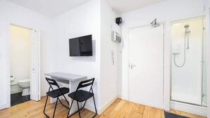 Ett litet rum med en vit dörr, en dusch, ett litet skrivbord med en stol och en tv som är monterad på väggen.