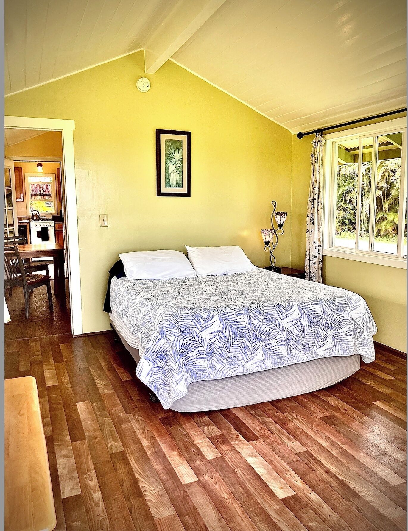 Ein Schlafzimmer mit Bett, Holzboden, einem Fenster mit Vorhängen, einem gerahmten Bild an der Wand und einem durchgehenden Durchgang erkennbaren Küchenbereich.
