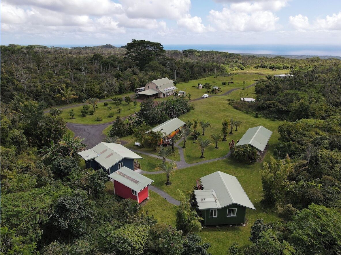 An aerial view of a property with several buildings surrounded by lush greenery and a clear view of the ocean.
