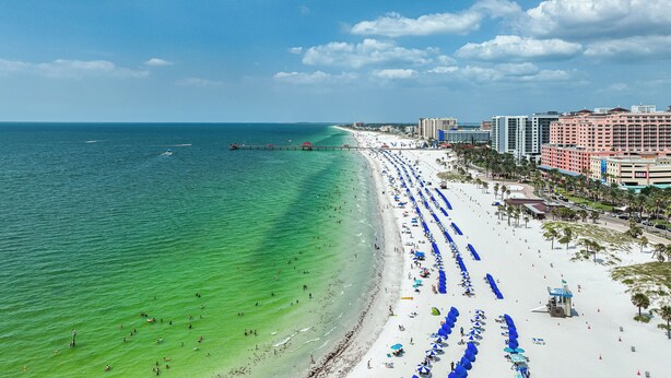 A beach with a long stretch of umbrellas and beach chairs, a pier extending into the water, and a row of hotels in the background. - Ocean Front Beach in Clearwater 2BB 2BA  (Clearwater Beach)