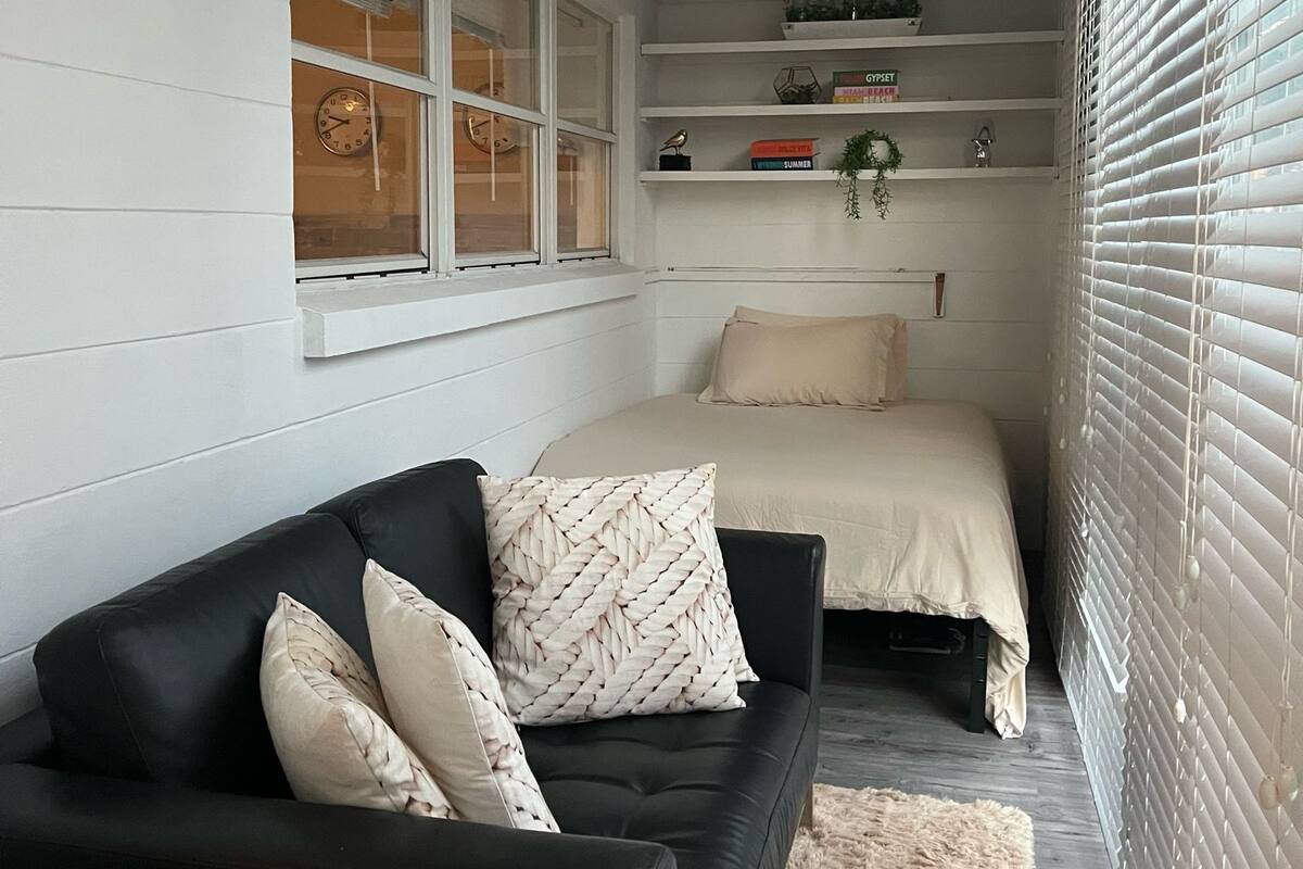 Un dormitorio con un sofá de cuero negro, una cama con cubrecolcha color beige, una ventana con un reloj y una estantería con objetos decorativos.