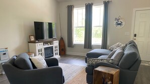 Een woonkamer met twee grijze banken, een houten salontafel, een flatscreen televisie op een kast, en een raam met gordijnen.