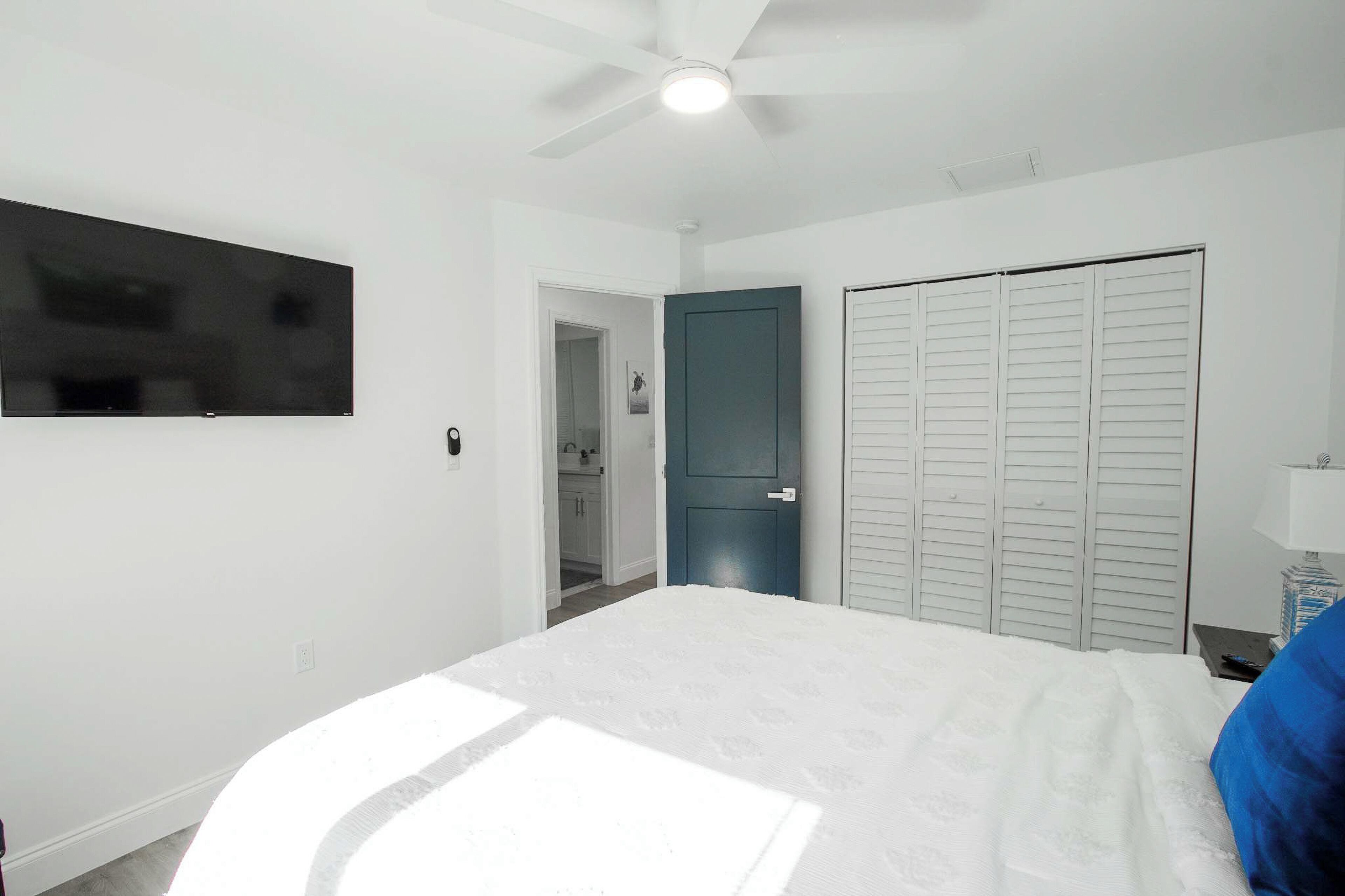 Un dormitorio con cama, ventilador de techo, televisor, armario y puerta al baño.