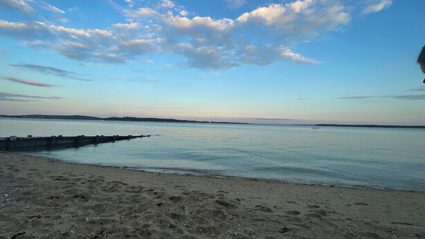 Una playa con un mar tranquilo, cielo despejado y una costa lejana.