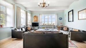 A spacious living room with a large black leather sofa, a wooden coffee table, and a chandelier. There are two windows with curtains, a television, and a cabinet. - Gorgeous home in Bredebro with WiFi (Bredebro)