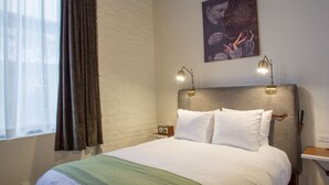 Une chambre d’hôtel avec un lit, une table de chevet, une lampe, un tableau au mur et une fenêtre avec des rideaux.