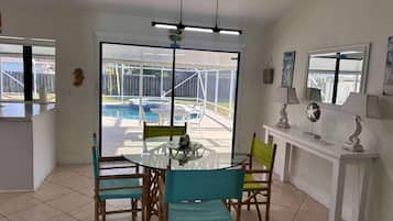 Un coin repas avec une table et des chaises en verre, une console blanche agrémentée de lampes, et une vue sur une piscine à travers de grandes portes vitrées.