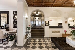 Eine Hotelhalle mit Schachbrettmuster-Boden, einer Treppe, einem Empfangstresen und einer Sitzecke.