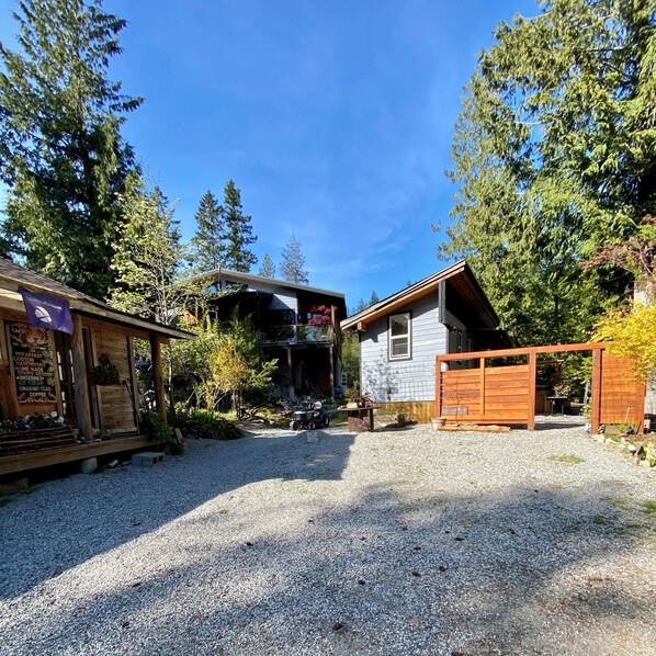 A rustic cabin with a gravel driveway, surrounded by tall trees and a wooden fence.