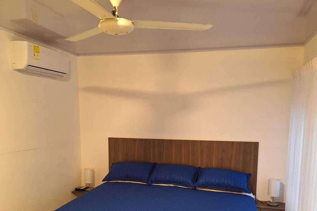 A hotel room with a large bed, a ceiling fan, an air conditioning unit, and a nightstand with a lamp.