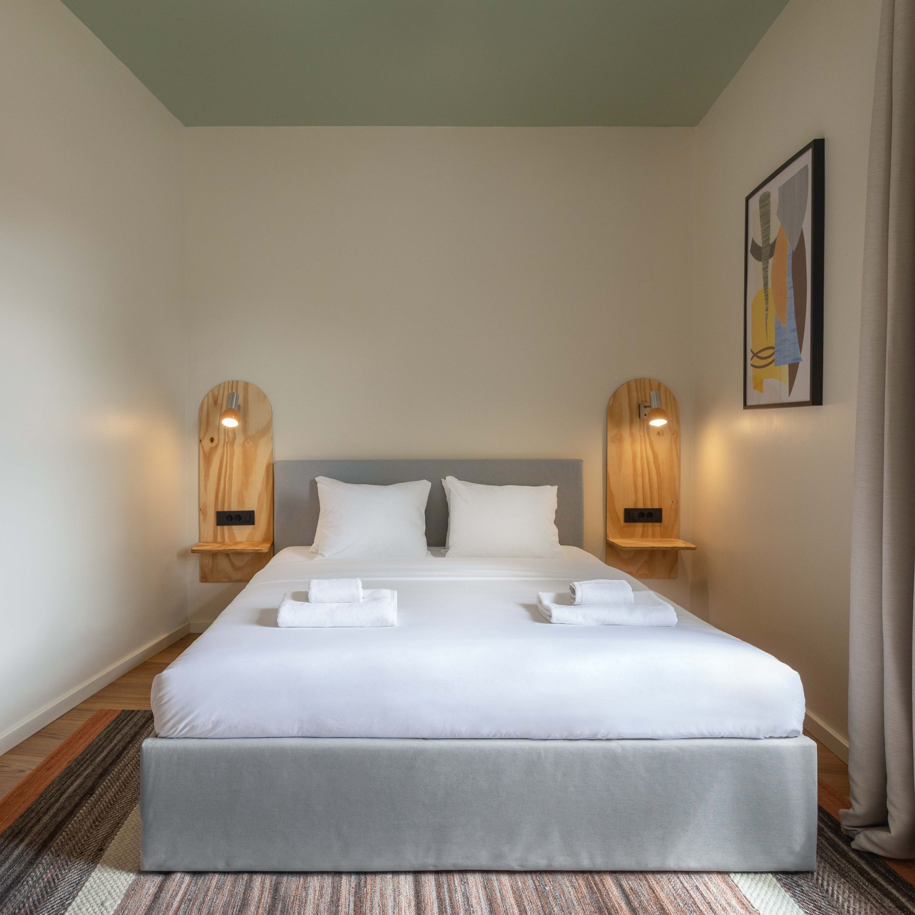 A neatly made bed with white linens and two pillows, flanked by wooden bedside tables with wall-mounted lamps.