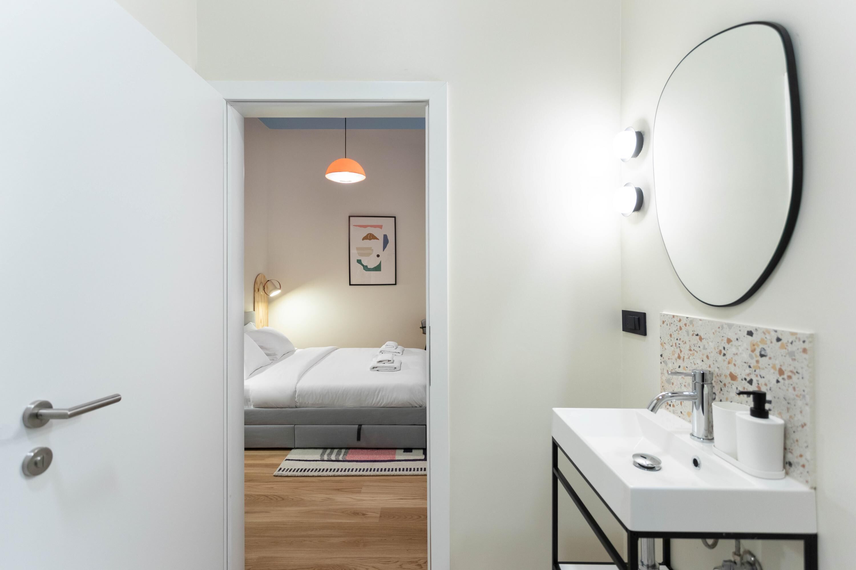 A modern hotel room with a white door, a bathroom with a large mirror and sink, a bed with a gray headboard, and a small rug.
