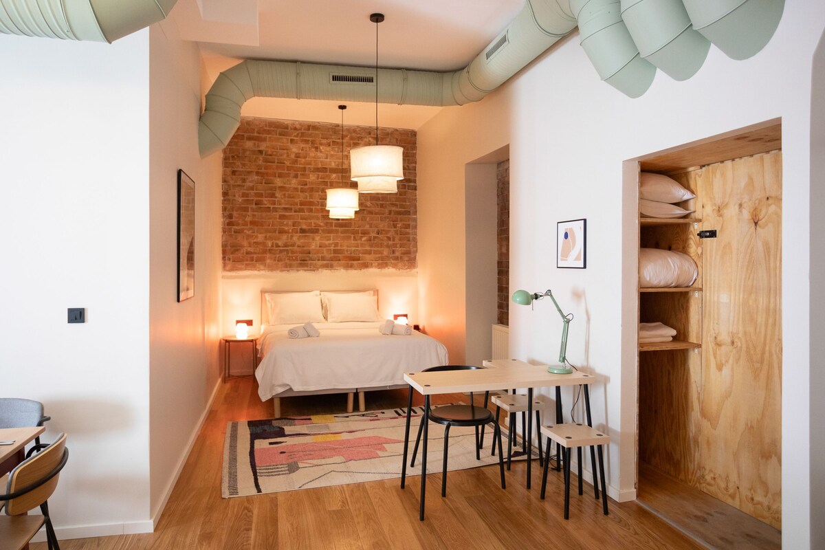 A cozy bedroom with a brick wall, a bed, bedside lamps, a desk with chairs, and a wooden wardrobe.