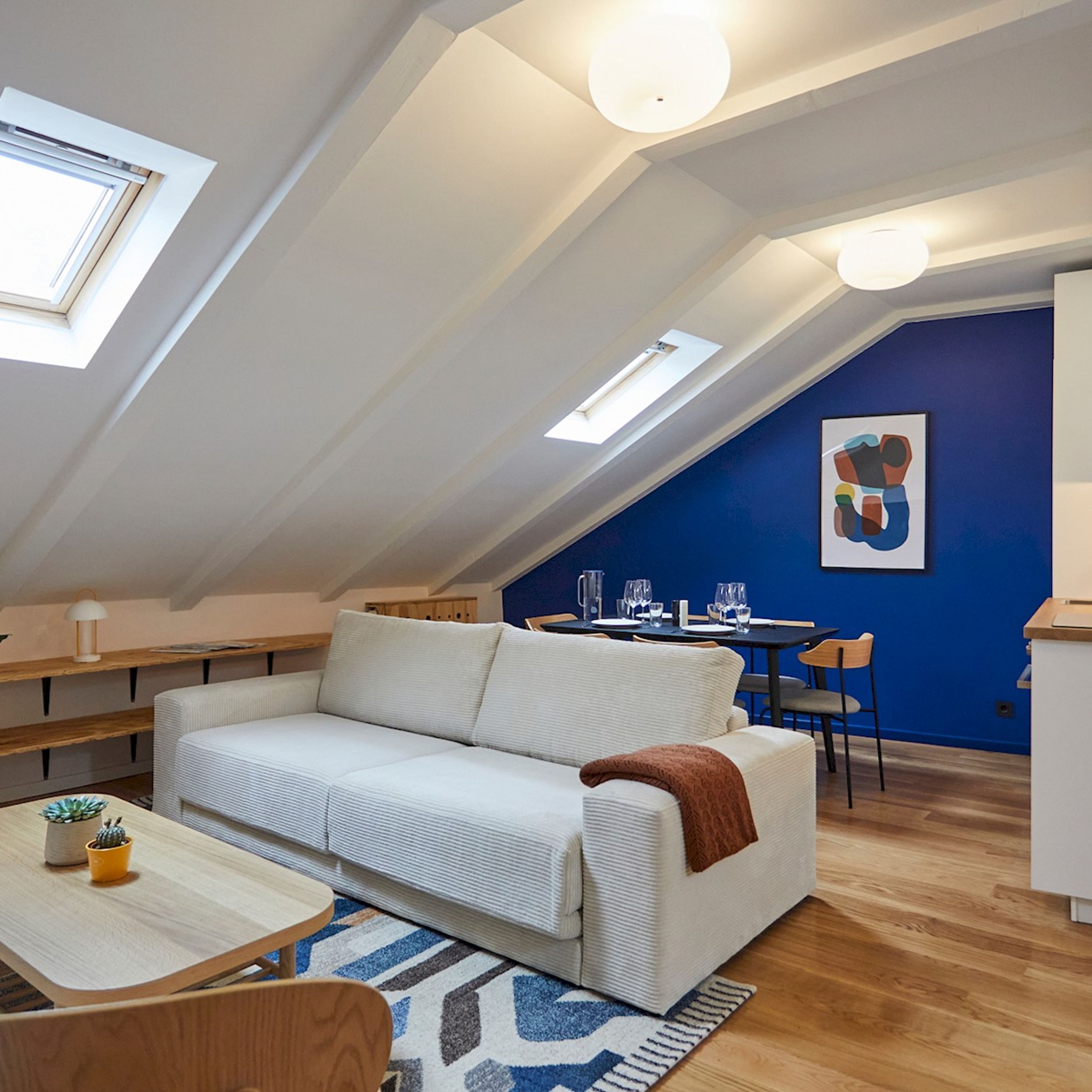 Un appartement-loft chaleureux avec un plafond en pente, un canapé blanc, une petite table à manger et un mur d’accent bleu.