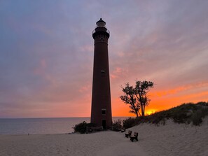 Ett fyrtorn med en fasad av tegel, beläget på en sandstrand med en bänk och ett träd i närheten.