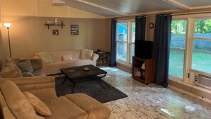 Ein Wohnzimmer mit beiger Couch, einem Couchtisch und einer TV-Konsole. Es gibt große Fenster mit Vorhängen, einen Kamin und eine Deckenleuchte.